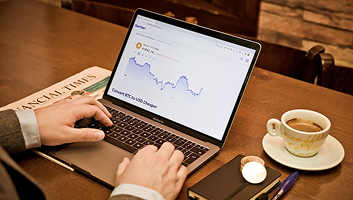 Hands typing on a laptop showing a financial chart on a desk with coffee and newspaper
